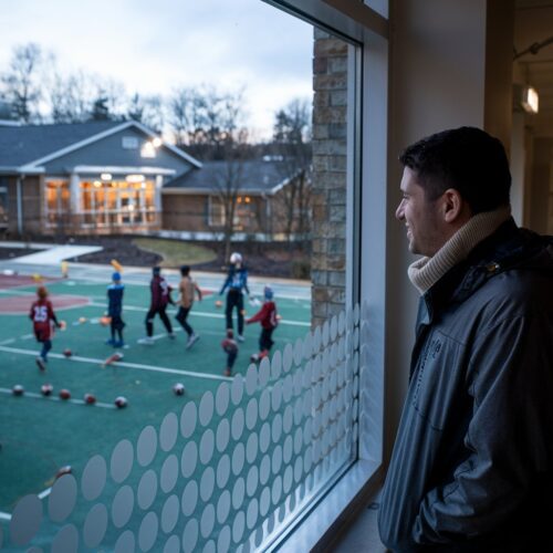 Mann beobachtet Kinder auf städtischem Ballspielplatz aus Fenster.