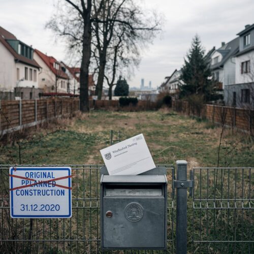 Unbebautes Grundstück in Münster mit übergrownem Gras, Mahnschreiben im Briefkasten, Baufristen überschritten.