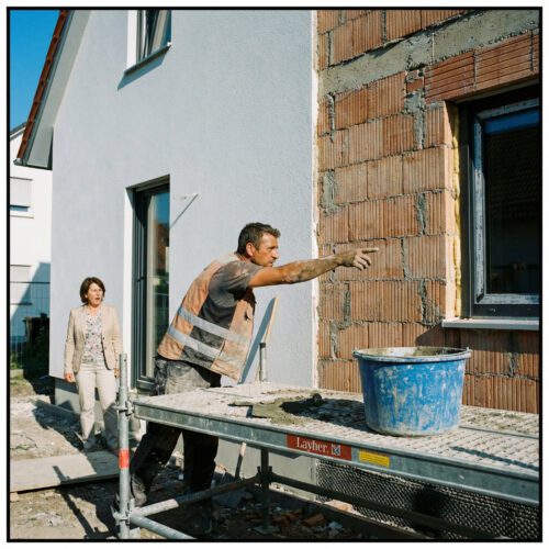 Ein Bauunternehmer zeigt mit fordernder Geste auf die abrupte Trennlinie der halb verputzten Hausfassade; die Bauherrin lauscht.