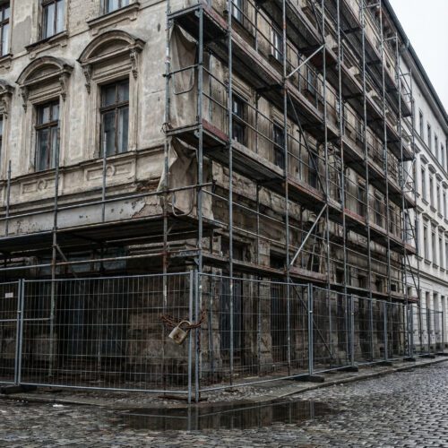 Ein massives Stahlgerüst umschließt vollständig die verlassene, verwitterte Fassade eines Altbaus.