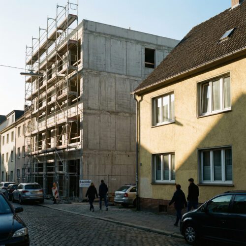 Ein massiver Neubau mit Gerüst wirft eine harte diagonale Schattenkante auf die helle Fensterfront eines Nachbarhauses.