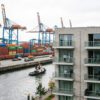 baugenehmigung-studierendenwohnheim-hafen-gemengelage Modernes Studierendenwohnheim mit Balkonen direkt neben einem Hafen-Containerterminal mit hohen Kränen und Frachtcontainern.