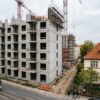 klage-baugenehmigung-nachbar-abstandsflaechen Massiver Rohbau eines Wohnblocks neben einem kleinen Wohnhaus, getrennt durch einen schmalen unbebauten Bodenstreifen.